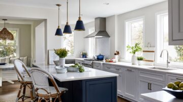 Modern kitchen with white cabinets, a large island with blue base, pendant lights, wicker bar stools, and windows letting in natural light. Fruits and plants decorate the counters.