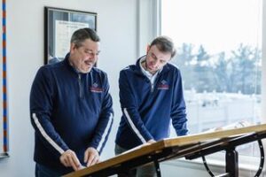 Two men in matching navy pullovers stand at a drafting table, demonstrating team work as they review documents together in a bright office with a window and framed certificate on the wall.