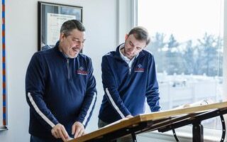 Two men in matching navy pullovers stand at a drafting table, demonstrating team work as they review documents together in a bright office with a window and framed certificate on the wall.